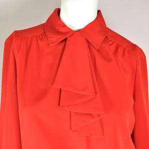 Könnte beinhalten: Eine leuchtend rote Bluse mit einem markanten Krawattendetail. Das langärmlige Hemd hat einen klassischen Kragen und eine kaskadenförmige, mehrschichtige Krawatte, die einen raffinierten und eleganten Look erzeugt. Die einfarbige Farbe und das Design eignen sich für verschiedene Anlässe.