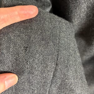 May include: Close-up of a dark gray herringbone fabric. The fabric has a subtle pattern and texture, with a visible seam. The image highlights the details of the material, showcasing its weave and color variations.