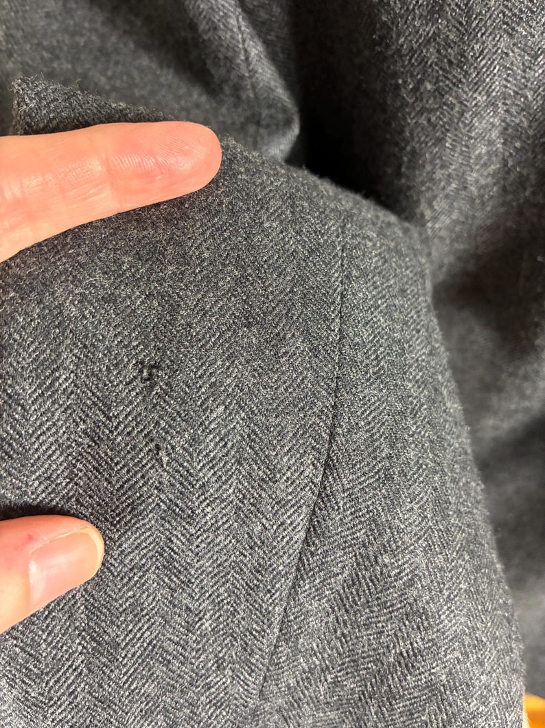 May include: Close-up of a dark gray herringbone fabric. The fabric has a subtle pattern and texture, with a visible seam. The image highlights the details of the material, showcasing its weave and color variations.