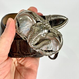 May include: A brown leather belt with a silver elephant-shaped buckle. The buckle features a detailed elephant head and decorative leaves. The belt has a textured surface and a classic design, suitable for various outfits.