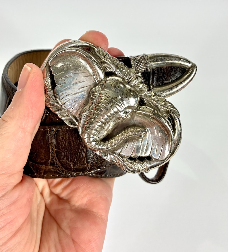 May include: A brown leather belt with a silver elephant-shaped buckle. The buckle features a detailed elephant head and decorative leaves. The belt has a textured surface and a classic design, suitable for various outfits.