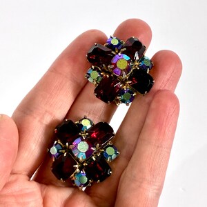 May include: Two vintage floral-shaped earrings. Each earring features a cluster of dark red, iridescent, and green crystals set in a gold-tone metal. The crystals are arranged in a flower-like pattern, with a central, larger stone.
