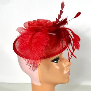 May include: A vibrant red fascinator with a circular base, adorned with feathers and a layered flower design. The headpiece features a mesh overlay and additional feather accents, creating a dramatic look. Perfect for special occasions.