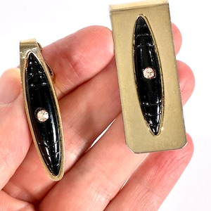 May include: A gold-coloured tie clip and money clip set. Each item has a black, oval-shaped centre with a textured pattern and a small, clear rhinestone. The clips are held in a hand against a white background.