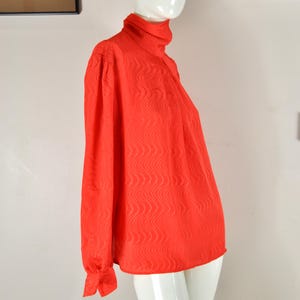 May include: A vibrant red long-sleeved blouse with a high neckline, displayed on a white mannequin. The blouse features a subtle wave pattern across the fabric. The sleeves are cuffed at the wrists, and the overall style is vintage.