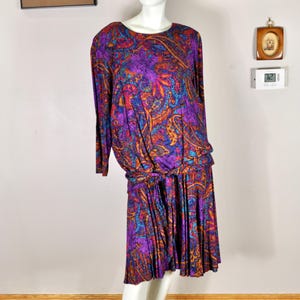 Może przedstawiać: Sukienka w stylu vintage na manekinie. Sukienka ma żywy wzór paisley w odcieniach fioletu, pomarańczy i niebieskiego. Ma długie rękawy, marszczoną talię i plisowaną spódnicę, prezentując styl retro.