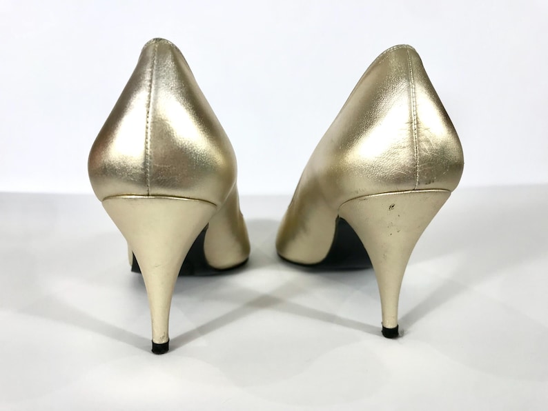 May include: A pair of gold-colored high heels with pointed toes and stiletto heels. The shoes have a metallic sheen and are set against a white background. The heels are approximately 3 inches tall.