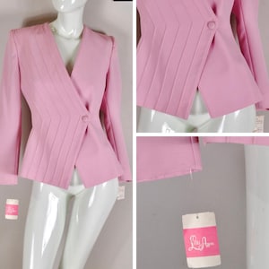 May include: A light pink blazer with a V-neck design and a single button closure. The blazer features pleated detailing on the front and long sleeves. A tag with the brand name "Lily Ann" is attached.