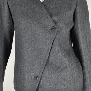 May include: A gray blazer with a wrap-style front and two dark buttons. The jacket has long sleeves and a V-neckline. The fabric appears to have a subtle herringbone pattern.