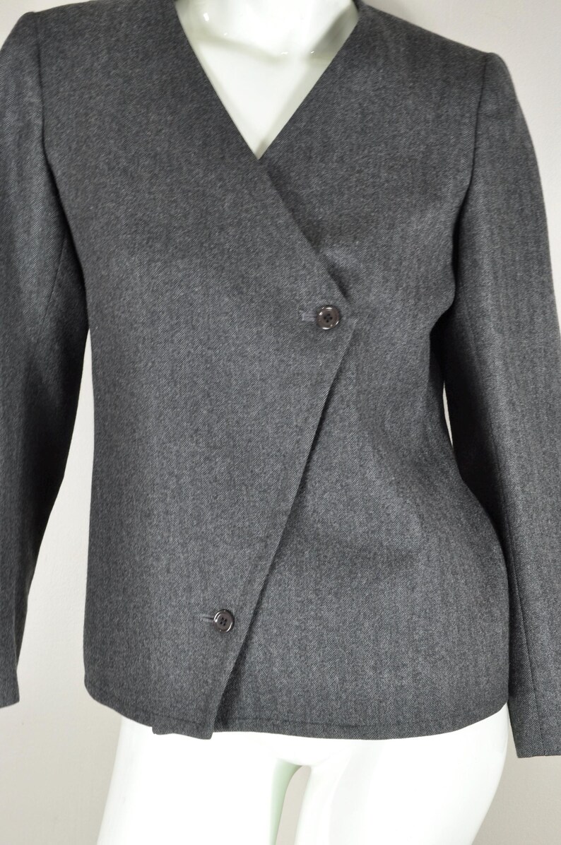 May include: A gray blazer with a wrap-style front and two dark buttons. The jacket has long sleeves and a V-neckline. The fabric appears to have a subtle herringbone pattern.