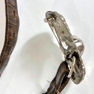 May include: Close-up of a brown leather belt with a silver-colored buckle. The belt has a textured surface and visible stitching. The buckle is ornate with a detailed design.