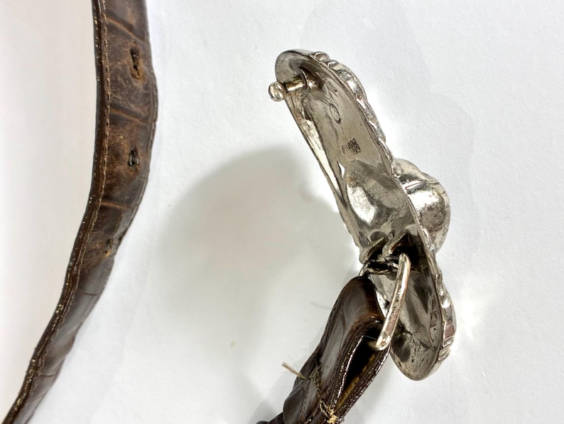 May include: Close-up of a brown leather belt with a silver-colored buckle. The belt has a textured surface and visible stitching. The buckle is ornate with a detailed design.