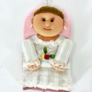 Puede incluir: Muñeca de peluche con forma de novia, con una cara redonda sonriente, pestañas oscuras y pendientes de perlas. La muñeca viste un vestido blanco con detalles de encaje y un adorno floral. Una parte trasera rosa y un velo completan el diseño.