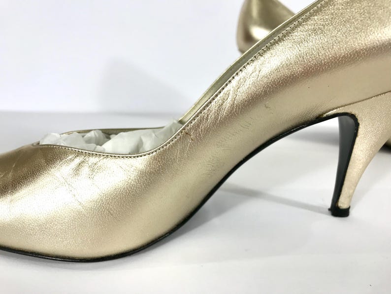 May include: Close-up of a pair of gold-colored high heels. The shoes have a pointed toe and a stiletto heel. The heels are approximately 3 inches high. The shoes are made of a shiny, metallic material.