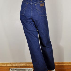 May include: Dark blue denim jeans with a high waist and a straight leg. The jeans feature a brown leather patch on the back pocket and a classic five-pocket design. The jeans are displayed on a mannequin.