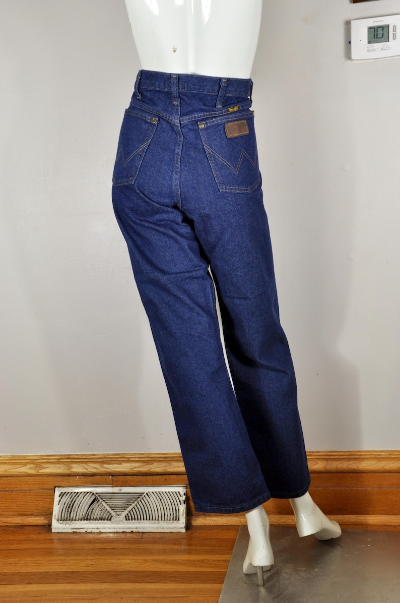 May include: Dark blue denim jeans with a high waist and a straight leg. The jeans feature a brown leather patch on the back pocket and a classic five-pocket design. The jeans are displayed on a mannequin.