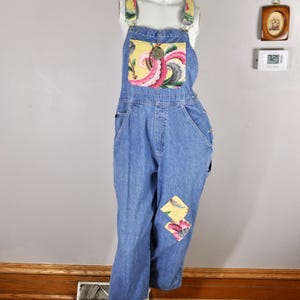 May include: Blue denim overalls with floral detailing on the bib, straps, and knee. The bib has a yellow background with pink and green floral patterns. A round, green button is on the bib. The overalls are displayed on a mannequin.