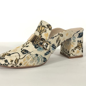 May include: White heeled mules with a floral pattern and a gold chain detail.