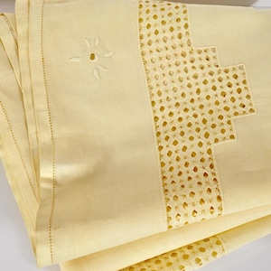 Puede incluir: Un tejido de color amarillo pálido que presenta una banda ancha de intrincado bordado calado con un patrón geométrico escalonado. La tela incluye un delicado motivo de ojal en forma de flor y un dobladillo recto con costuras detalladas.