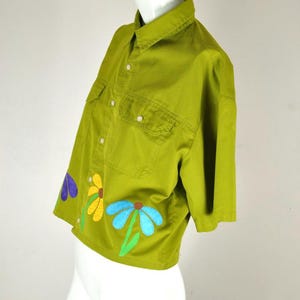May include: A short-sleeved, olive green button-down shirt featuring a collar, two chest pockets with white buttons, and a row of white buttons down the front. The bottom hem is decorated with three embroidered flower appliqués in purple, yellow, and blue.