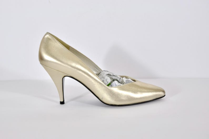 May include: Gold-colored high-heeled shoe with a pointed toe. The heel is slender and black-tipped. The shoe is filled with white paper. The shoe is likely made of leather or a similar material.