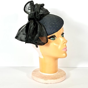May include: A navy blue hat with a polka dot pattern and a large black bow made of sheer fabric. The hat is displayed on a mannequin head with a pale complexion and green eyes. The hat is a vintage style.