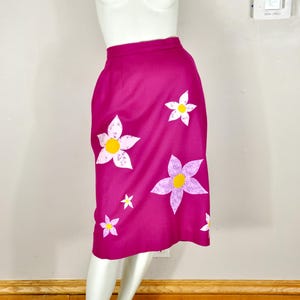 May include: A magenta skirt adorned with white and lavender flower appliqués with yellow centers. The skirt is displayed on a mannequin, showcasing its knee-length design. The floral pattern adds a whimsical touch to the garment.
