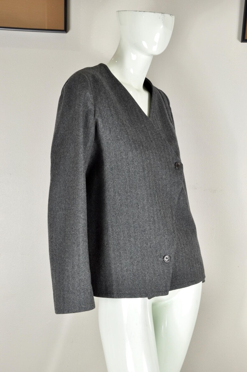 May include: A gray blazer with a V-neck and two dark buttons. The jacket is displayed on a white mannequin. The fabric has a subtle vertical stripe pattern. The blazer has long sleeves.
