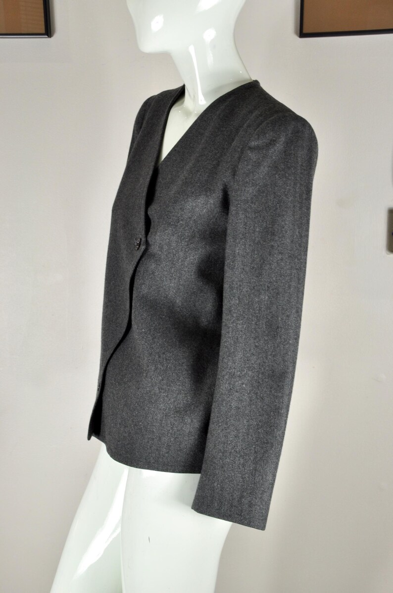 May include: A gray blazer with a V-neck and a single button closure. The jacket has long sleeves and a tailored fit. The fabric appears to be a wool blend. The blazer is displayed on a white mannequin.