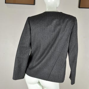 May include: A dark gray blazer with long sleeves, displayed on a white mannequin. The blazer has a simple, tailored design with a diagonal seam detail on the back. The fabric appears to be a wool blend.