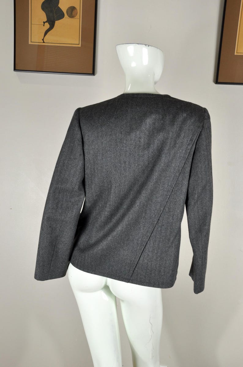 May include: A dark gray blazer with long sleeves, displayed on a white mannequin. The blazer has a simple, tailored design with a diagonal seam detail on the back. The fabric appears to be a wool blend.