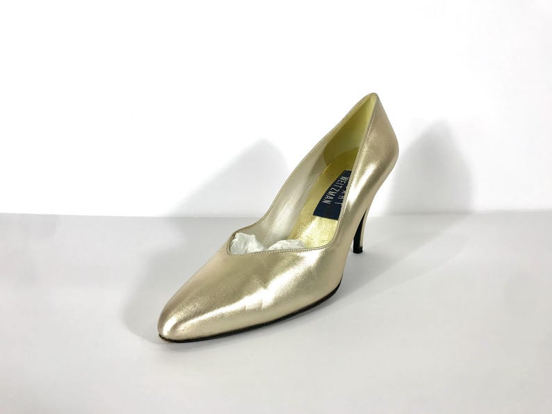 May include: A gold-colored high heel pump with a pointed toe. The shoe has a stiletto heel and a low-cut vamp. The brand name "Stuart Weitzman" is visible on the insole. The shoe is set against a white background.