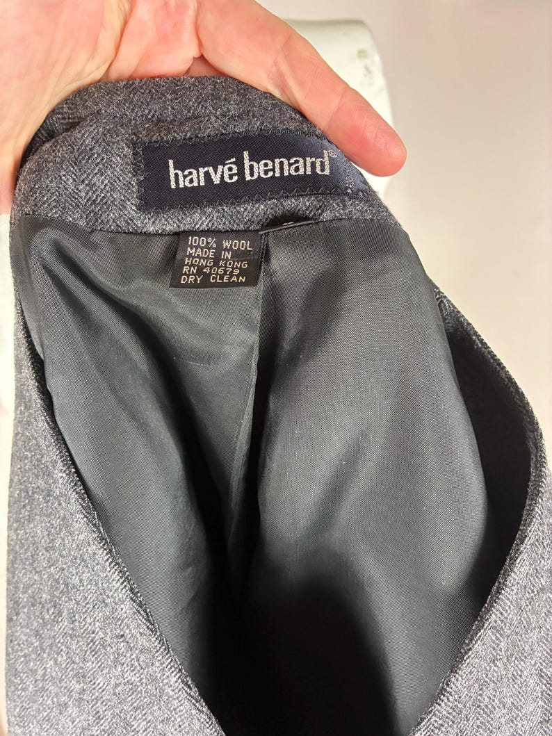 May include: Close-up of a gray wool blazer interior. The label reads "harv&eacute; benard" in white text on a black background. The tag states "100% wool, made in Hong Kong, RN 40679, dry clean."