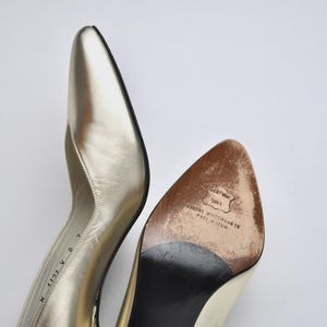 May include: A pair of gold-colored leather high heels with pointed toes. The sole of one shoe is visible, showing wear and the brand name "Stuart Weitzman Co. Made in Spain."