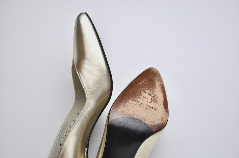 May include: A pair of gold-colored leather high heels with pointed toes. The sole of one shoe is visible, showing wear and the brand name "Stuart Weitzman Co. Made in Spain."