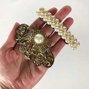 Puede incluir: Dos pinzas para el pelo, una con un diseño dorado adornado y una perla grande en el centro, y la otra con una fila de perlas blancas.