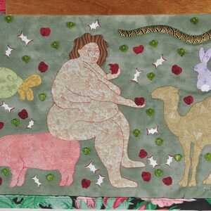 May include: A green fabric artwork depicting a nude woman sitting on a pink pig, holding an apple in one hand and a red heart in the other. A camel, a turtle, a rabbit, and a snake are also depicted in the artwork. The background is filled with green leaves, red hearts, and white stars.