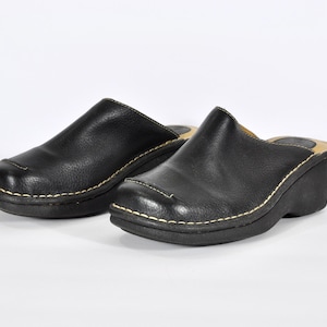 May include: Pair of black leather clogs with a rounded toe and a small heel. The clogs have white stitching around the sole and a smooth, matte finish. The shoes are set against a white background.