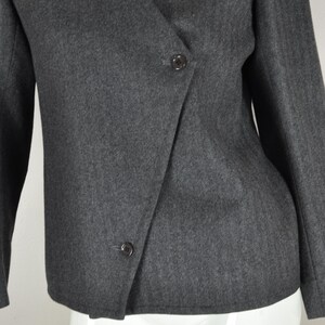 May include: A charcoal gray blazer with a wrap-style front and two dark buttons. The jacket has long sleeves and a V-neckline. The fabric appears to be a woven material.