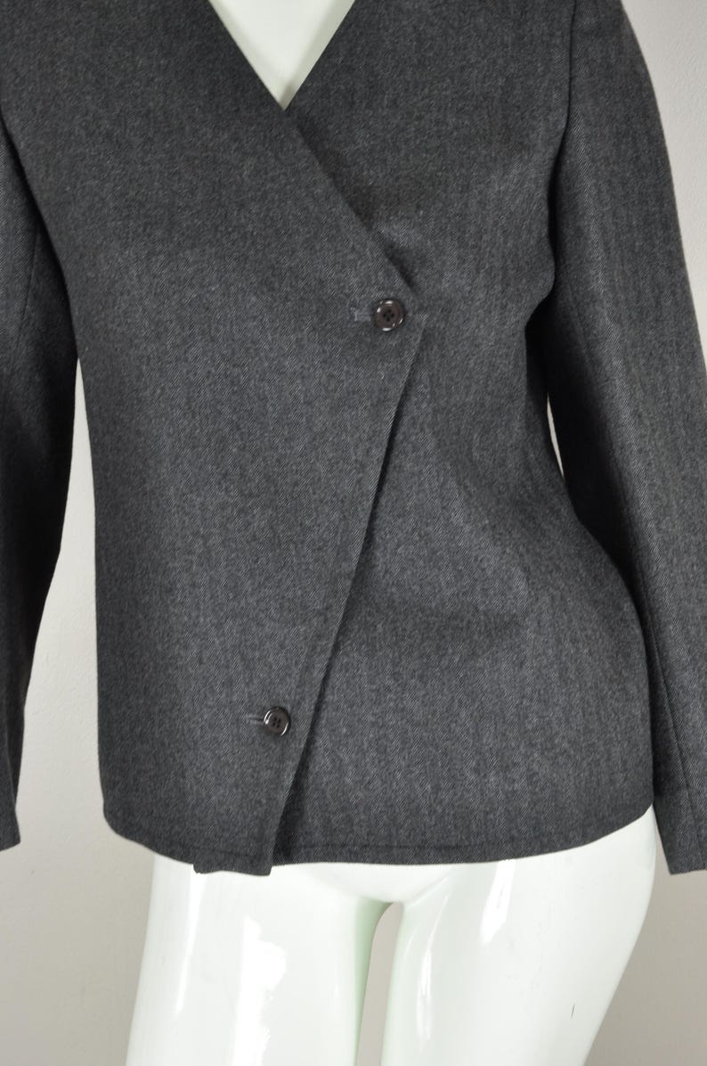May include: A charcoal gray blazer with a wrap-style front and two dark buttons. The jacket has long sleeves and a V-neckline. The fabric appears to be a woven material.