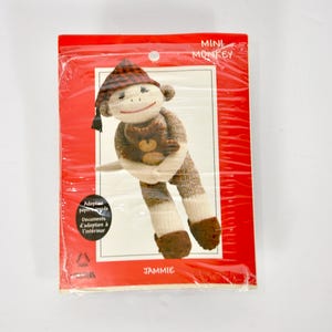 Puede incluir: Un juguete de mono de calcetín Mini Monkey llamado Jammie, empaquetado en una bolsa de plástico roja. El mono es marrón y crema con un gorro de cuadros rojos y negros, sosteniendo un pequeño oso de peluche marrón. El texto incluye Mini Monkey y Jammie.