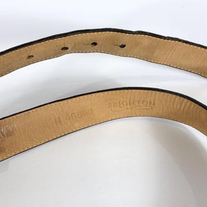 May include: A brown leather belt with a textured pattern. The inside of the belt is tan with the brand name "BRIGHTON" and the number "H 46850" printed on it. The belt has a silver-colored buckle.