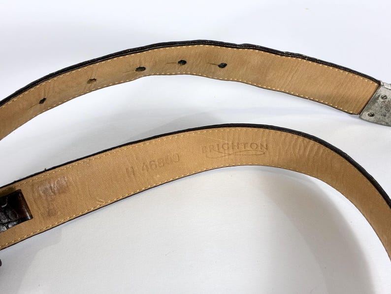 May include: A brown leather belt with a textured pattern. The inside of the belt is tan with the brand name "BRIGHTON" and the number "H 46850" printed on it. The belt has a silver-colored buckle.