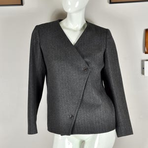 May include: A dark gray blazer with a V-neck and long sleeves. The blazer features a wrap-style front with two buttons for closure. The garment is displayed on a white mannequin.