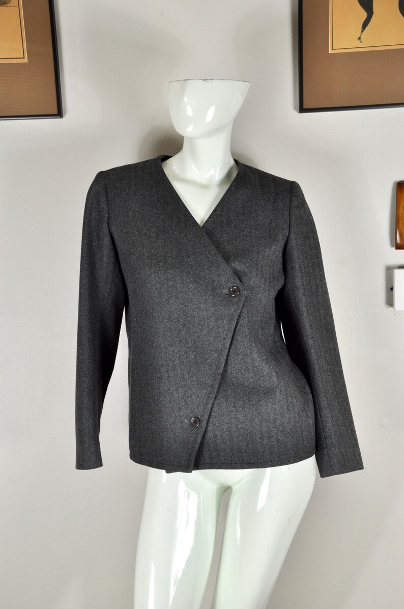 May include: A dark gray blazer with a V-neck and long sleeves. The blazer features a wrap-style front with two buttons for closure. The garment is displayed on a white mannequin.