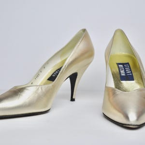 May include: A pair of gold-colored high-heeled pumps. The shoes have pointed toes and slender black heels. The interior of the shoes features a label with the brand name "Stuart Weitzman".