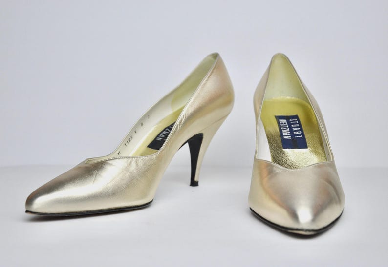 May include: A pair of gold-colored high-heeled pumps. The shoes have pointed toes and slender black heels. The interior of the shoes features a label with the brand name "Stuart Weitzman".