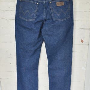May include: Dark blue denim jeans with the Wrangler logo on the back pocket. The jeans are hanging on a black hanger against a white brick wall. The jeans have a classic five-pocket design.