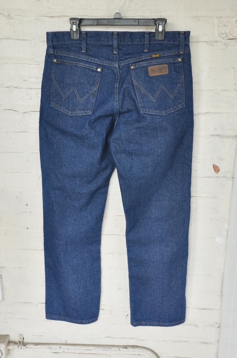 May include: Dark blue denim jeans with the Wrangler logo on the back pocket. The jeans are hanging on a black hanger against a white brick wall. The jeans have a classic five-pocket design.