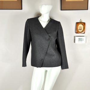 May include: A dark gray blazer with a V-neck and long sleeves. The blazer has a wrap-style front with two buttons. The fabric has a subtle herringbone pattern. The blazer is displayed on a white mannequin.